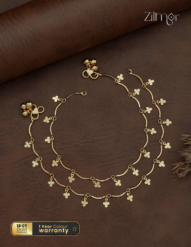 Zilmor 18K gold plated anklet pair with curved links, dangling clover-shaped charms and small bell-shaped accents.